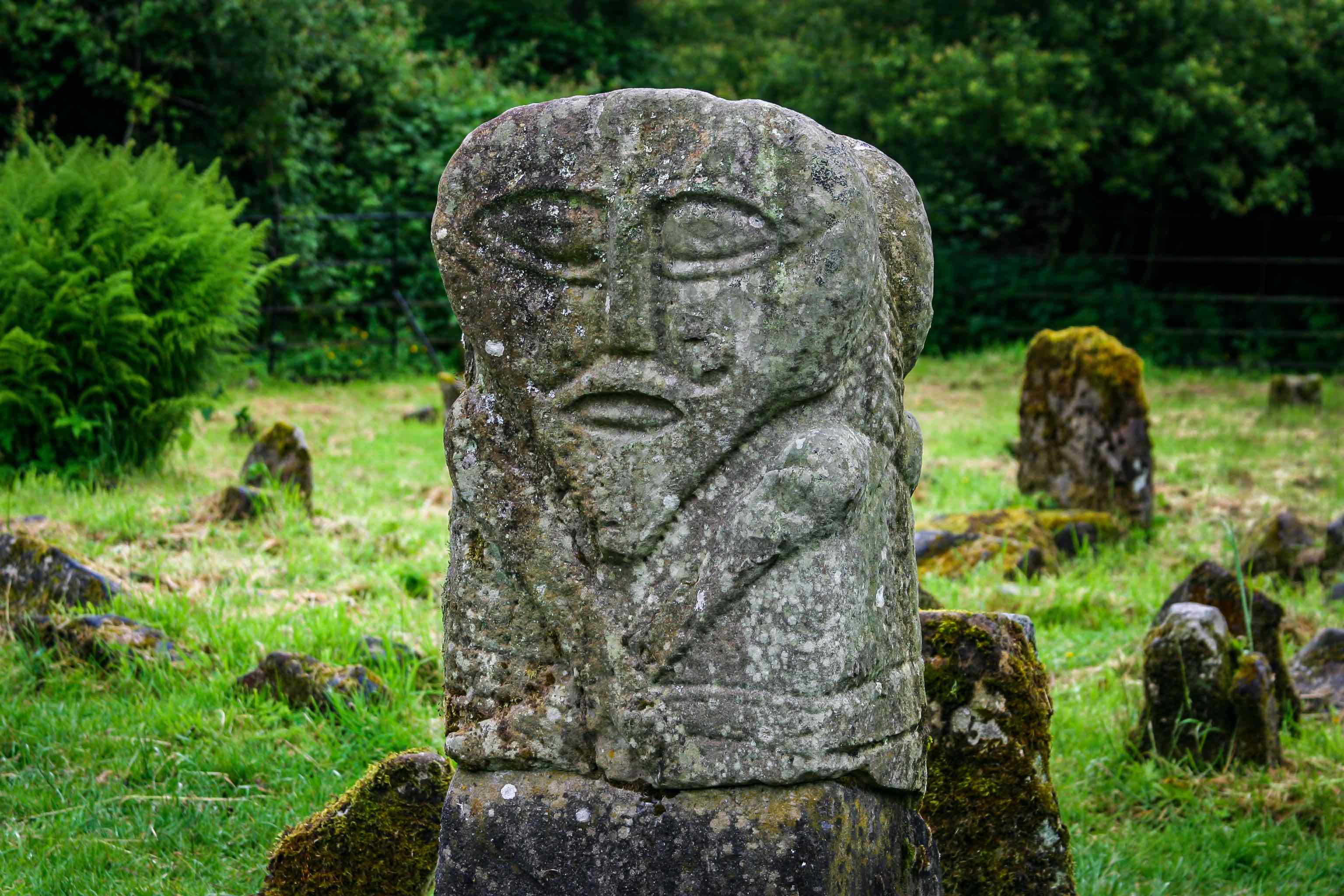 Close-up of ancient carved Janus stone figure with weathered features set among grass and moss-covered stones on Boa Island.