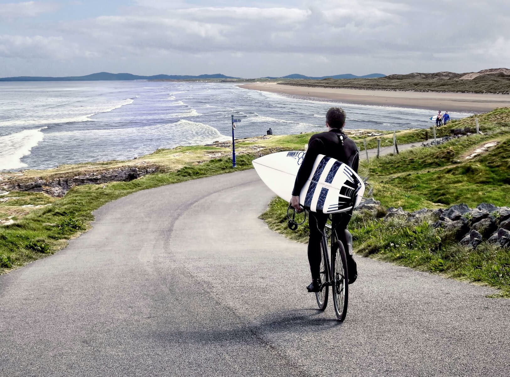 Surfer carrying a board while cycling along coastal path overlooking Bundoran beach and Atlantic waves.