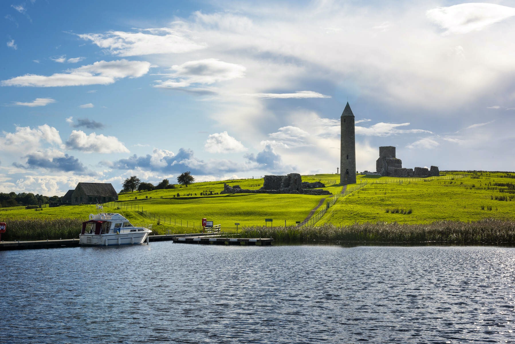 Boat docked at Devenish Island near medieval ruins and tall stone round tower set against rolling green fields and sky.