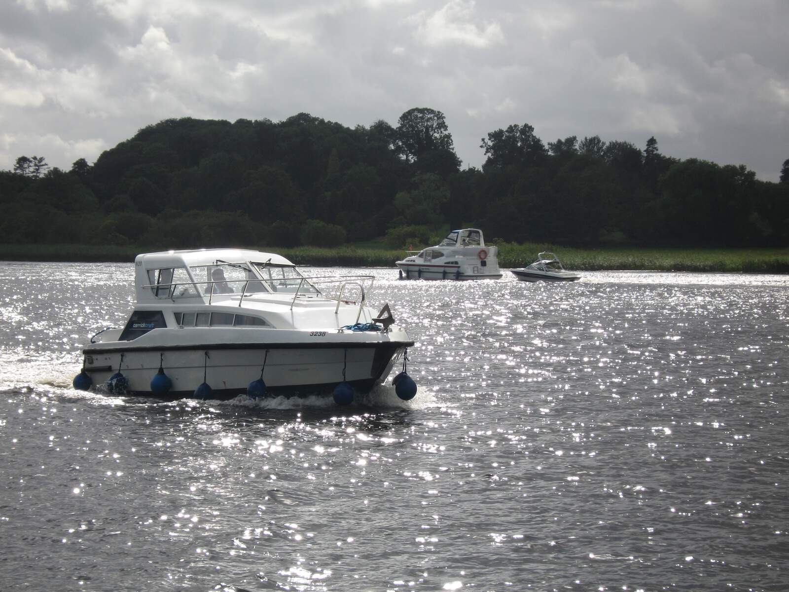 White motorboat cruising across Lough Erne waters with more boats behind and forested hills under cloudy skies.