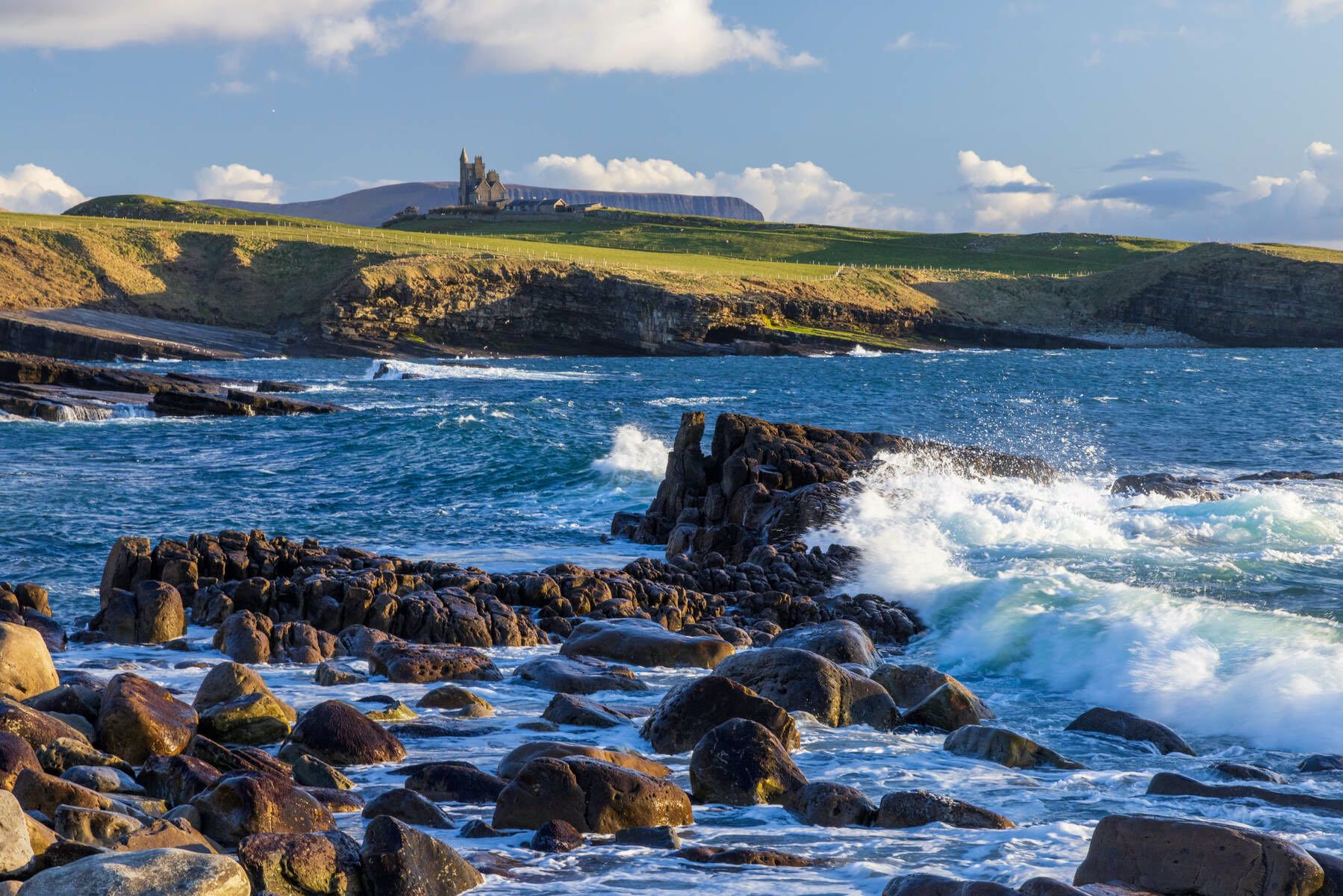 Rocky Atlantic shoreline at Mullaghmore with waves crashing and Classiebawn Castle visible on the distant headland.