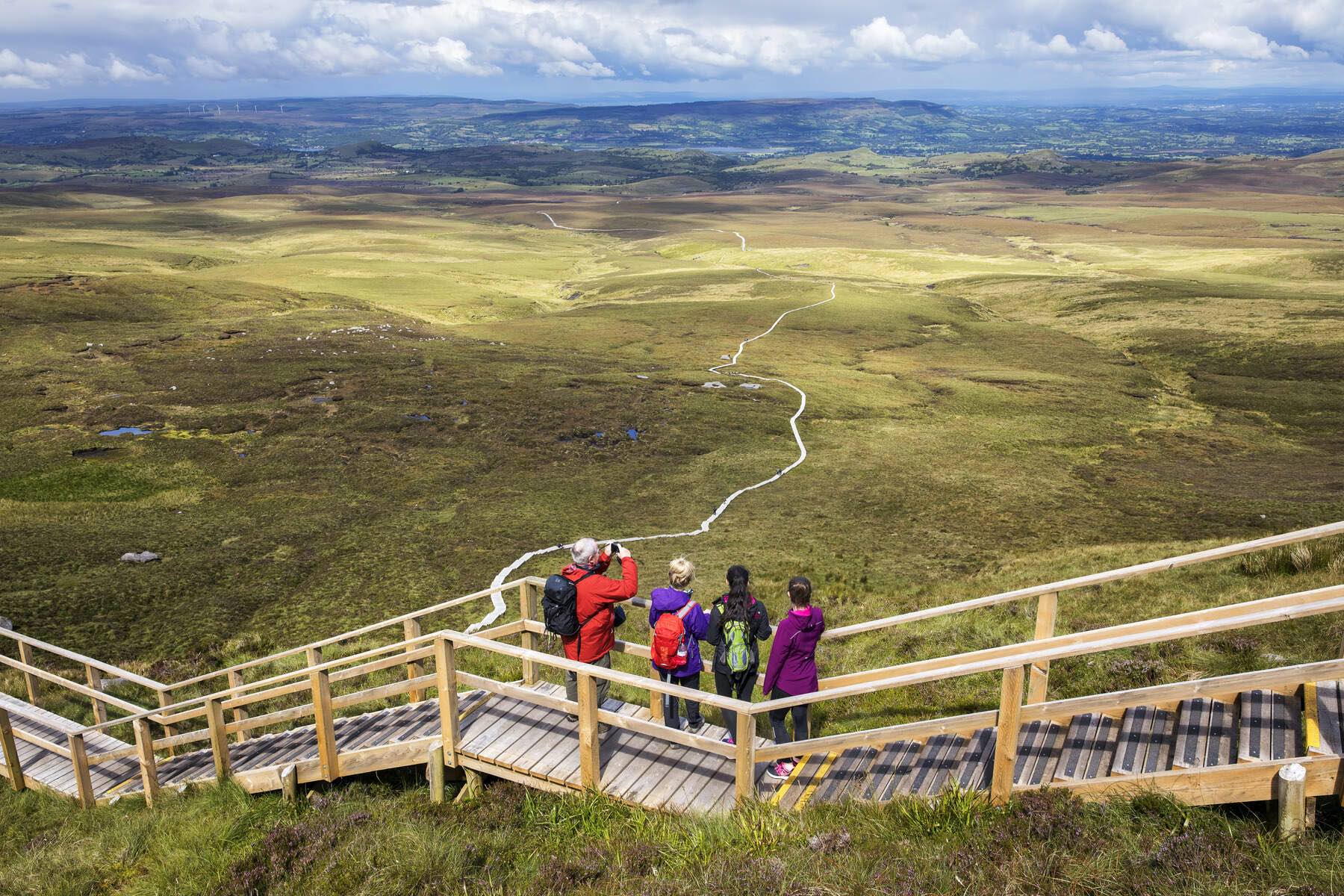 Group of hikers stand on wooden steps looking over rolling moorland with the winding Cuilcagh Boardwalk below.