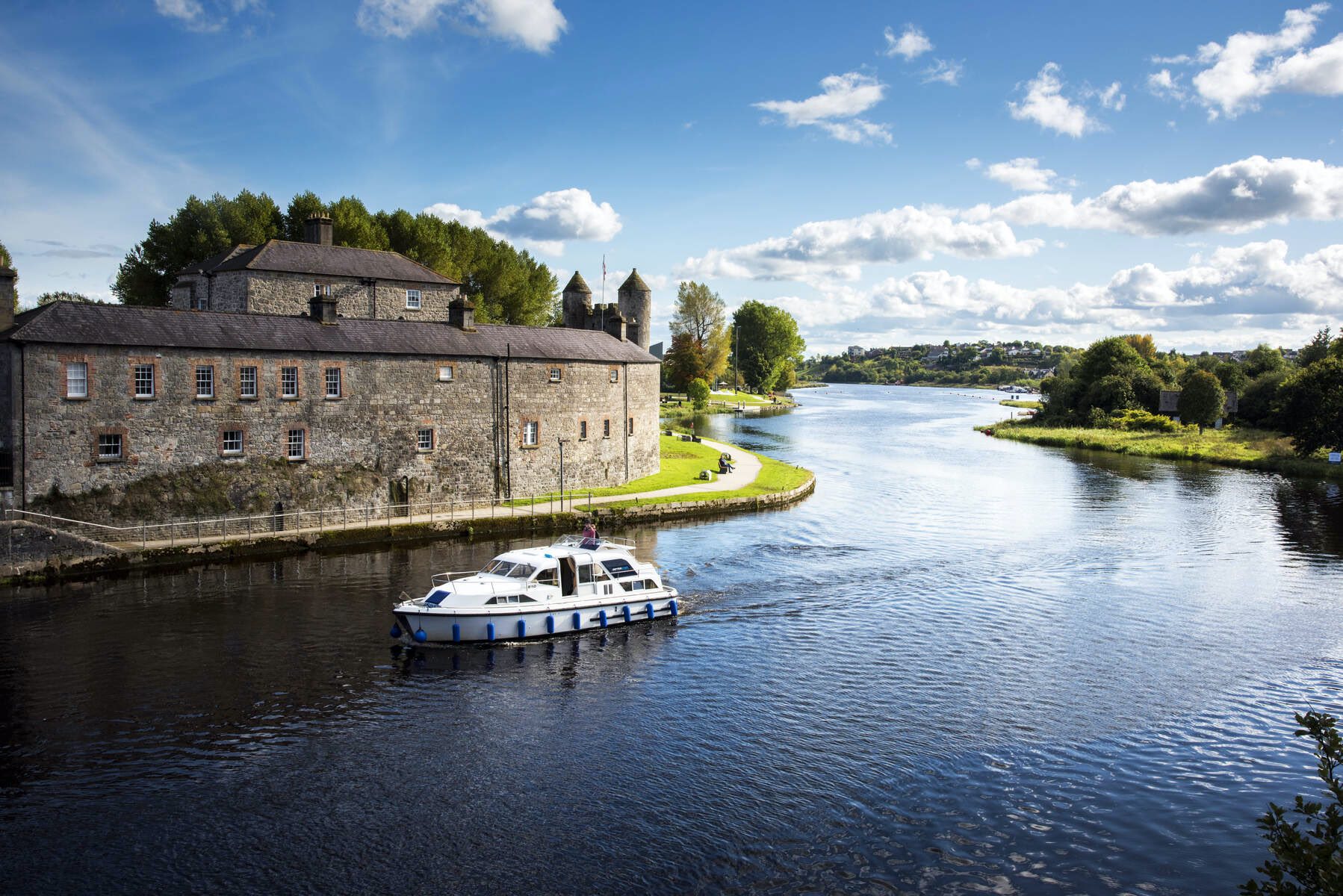 Boat cruising past Enniskillen Castle along the River Erne with stone walls, turrets, and green banks on a sunny day.