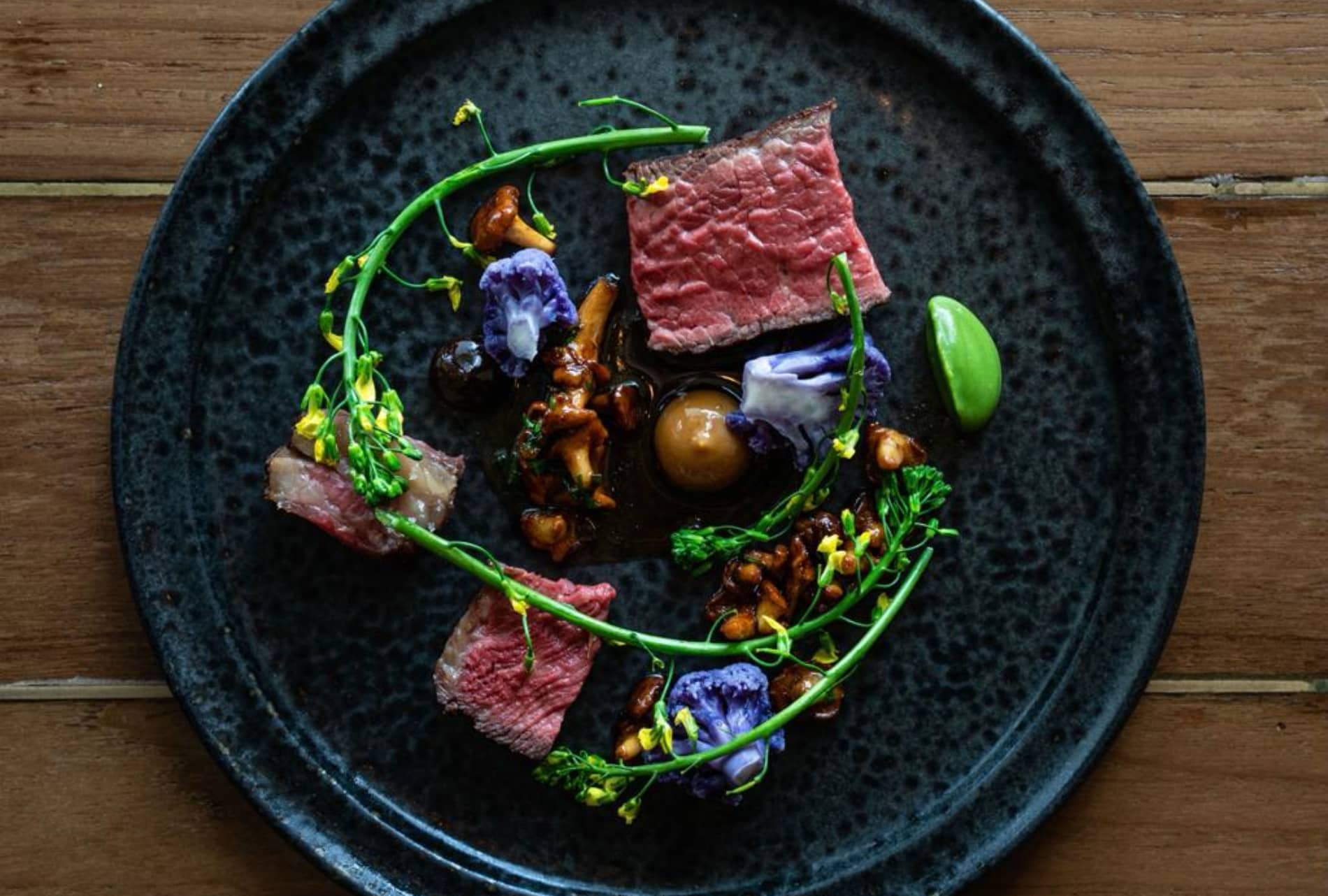 Artful dish of beef, chanterelles, broccoli and purple cauliflower garnished with herbs on a dark plate.