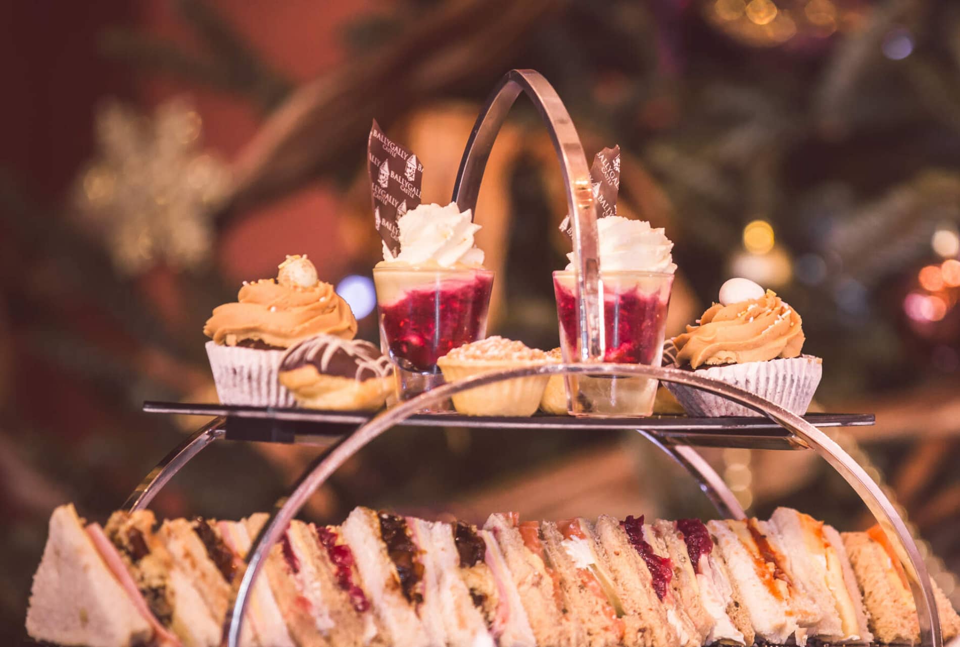 Festive tray with cupcakes, tarts, sandwiches, cream-topped desserts, and warm Christmas lights.