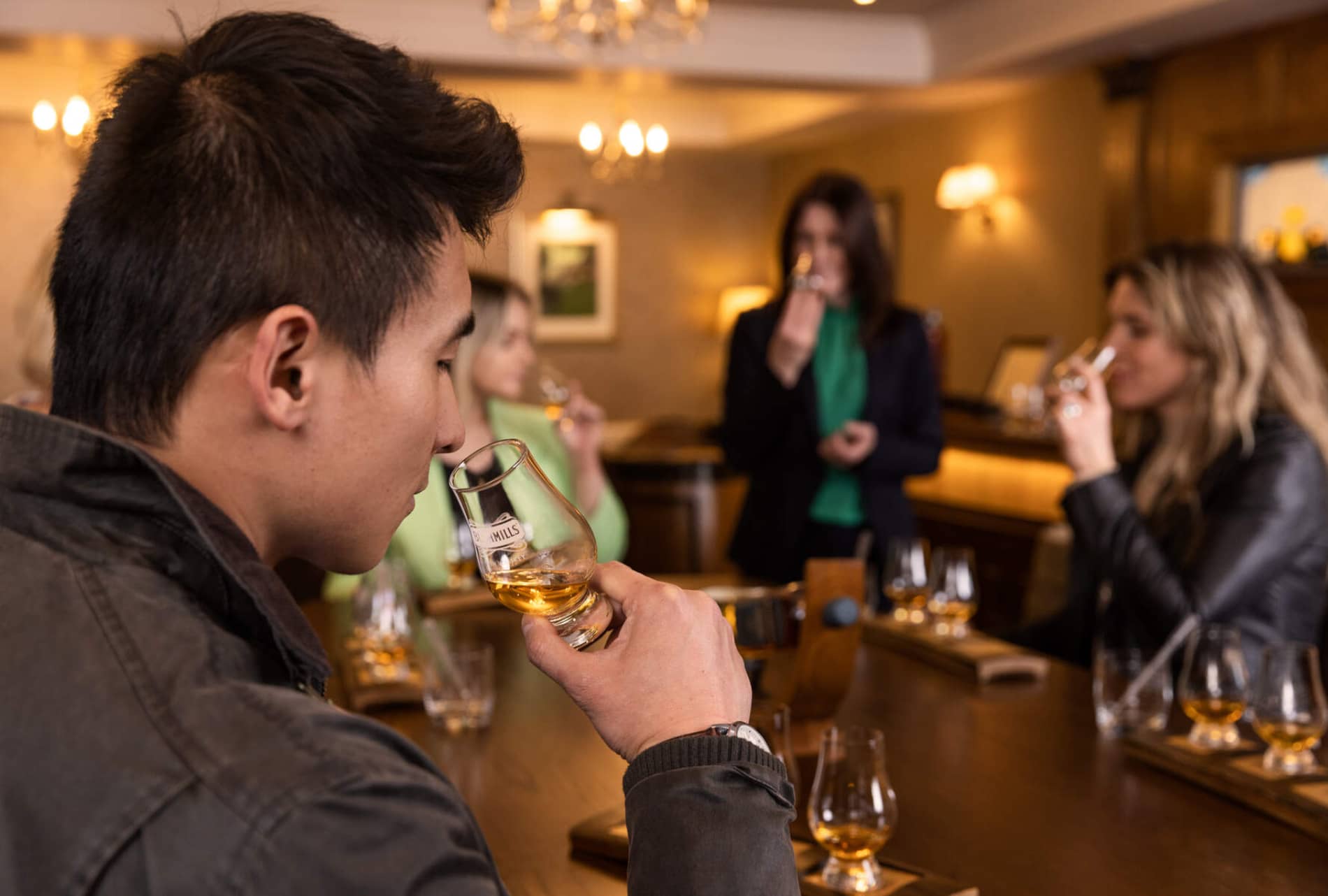 Visitors sample whiskey in a warm tasting room, glasses raised as they nose aromas and sip gently.