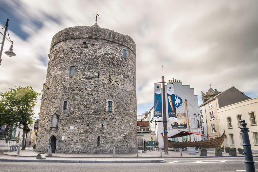 Round stone tower in Waterford city centre, with a replica Viking ship and flags nearby.