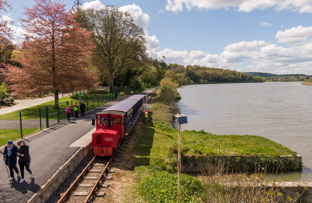 Small red train travelling along tracks beside a riverbank in County Waterford with walkers nearby.
