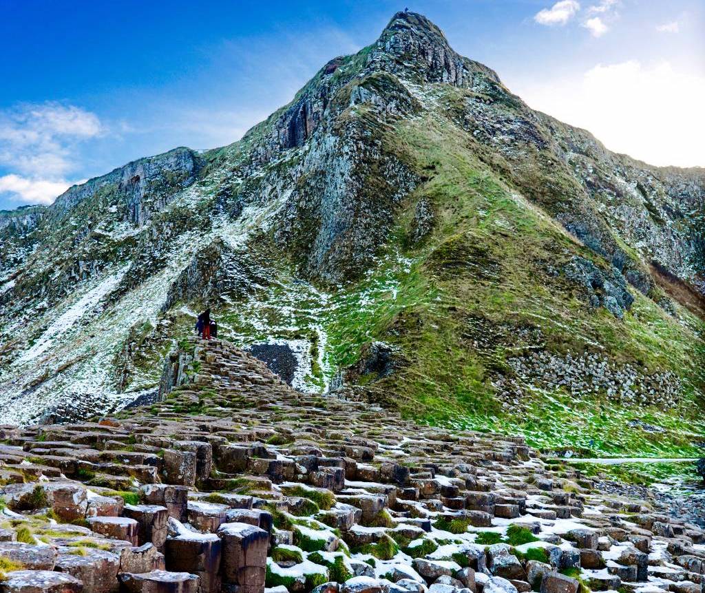 Snow-dusted basalt columns of the Giant’s Causeway rising toward a steep green cliff under a bright blue winter sky.