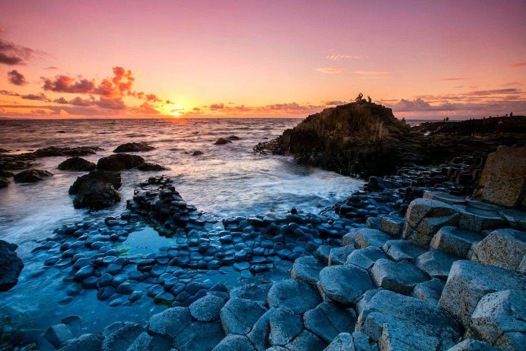 Giant’s Causeway at dusk with crashing waves, basalt columns, and vivid pink and orange sky as the sun sets.