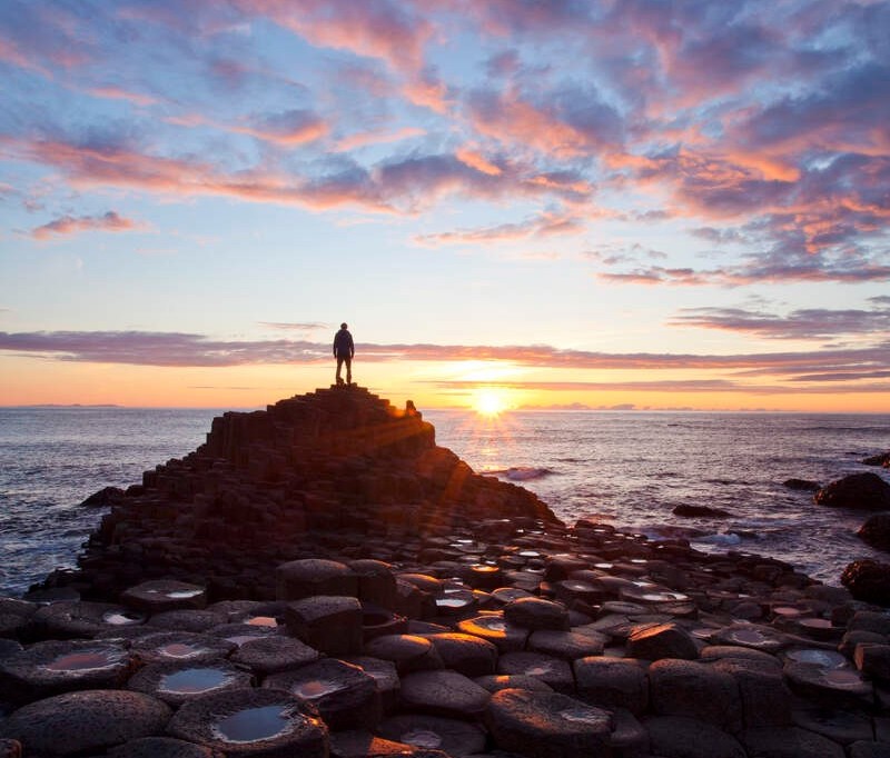 Person silhouetted on Giant’s Causeway columns at sunset, with golden light reflecting on wet basalt stones and sea.
