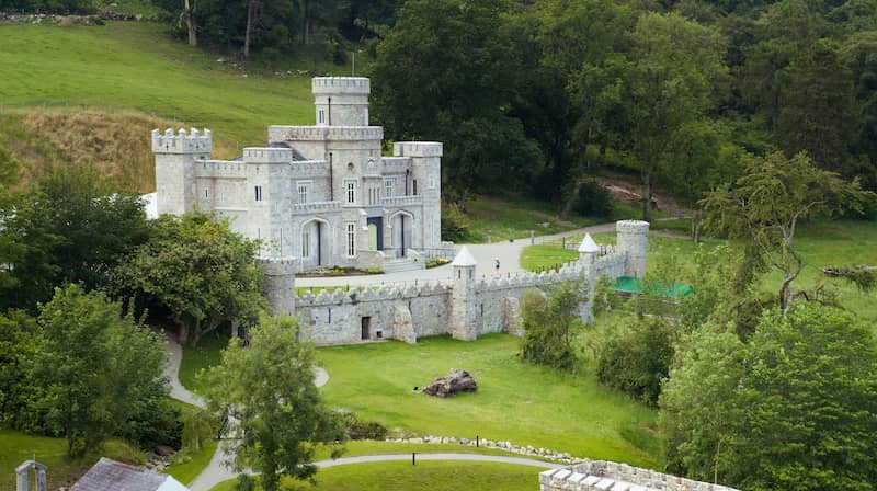 Small turreted castle with arched gates and high stone walls nestled in green countryside.