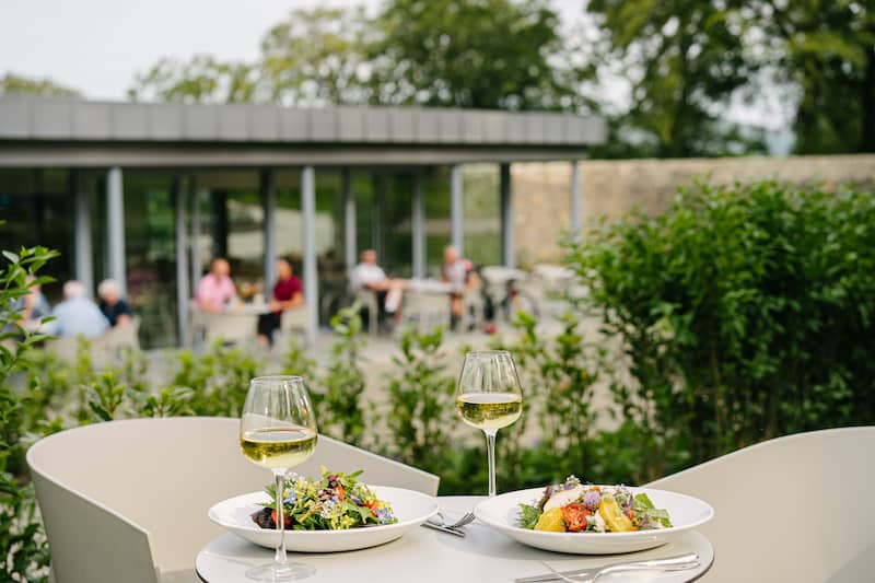 Two plates of salad and glasses of white wine set on a table outdoors, with people chatting behind.