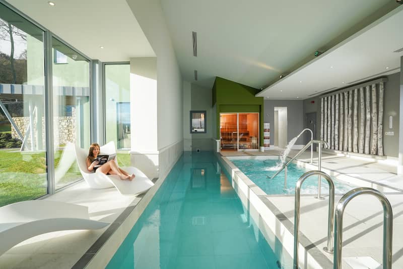 Modern indoor spa with loungers, a small pool, water jets and floor-to-ceiling windows.