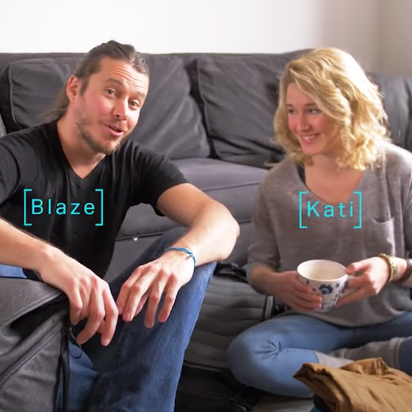 A man and woman seated on the floor beside a grey sofa, chatting over coffee in a cosy indoor setting.