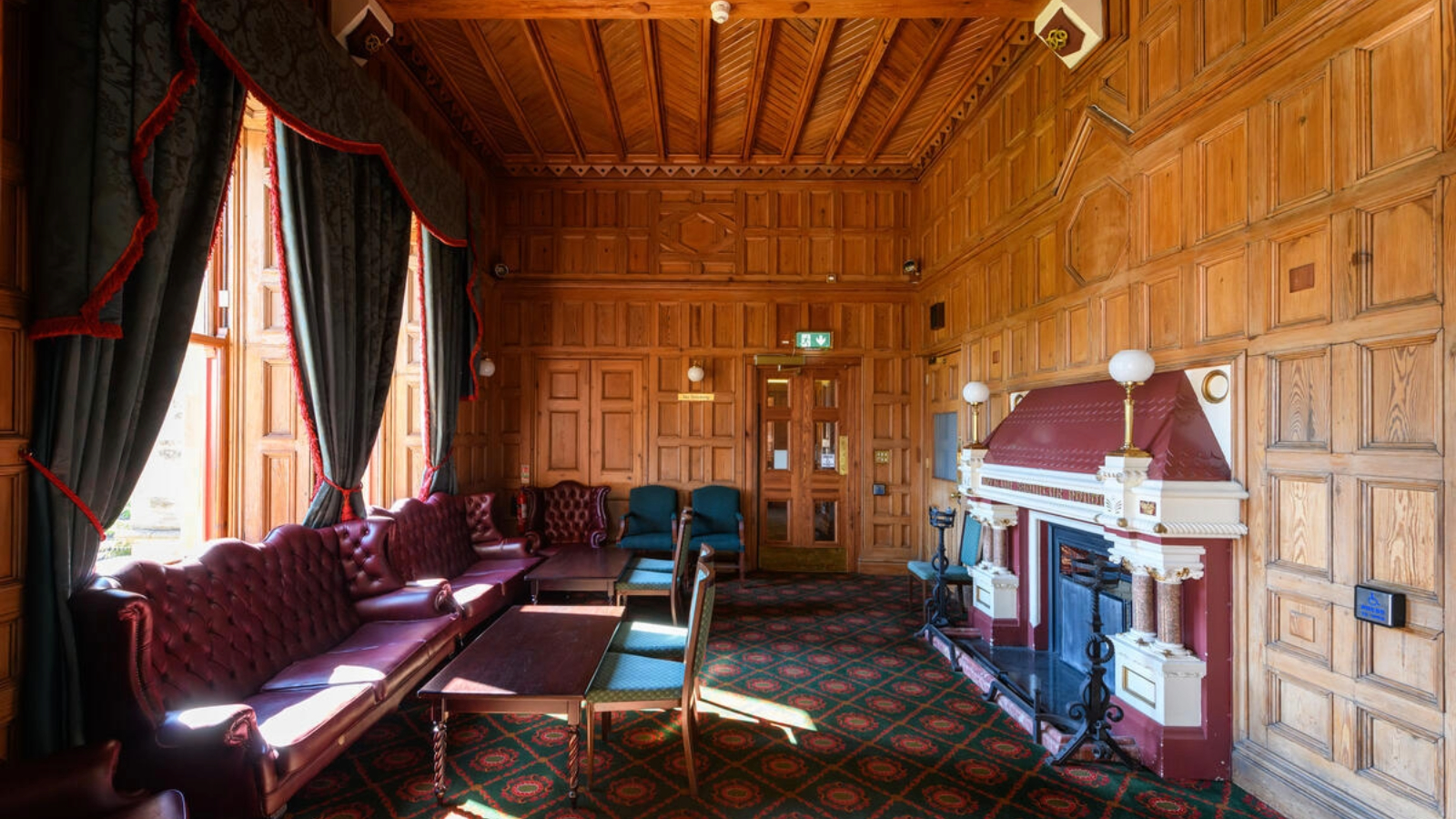 Ein holzvertäfeltes Wohnzimmer im Belfast Castle mit einem kunstvollen Kamin, Ledersofas und hohen Fenstern mit Blick auf das Gelände.