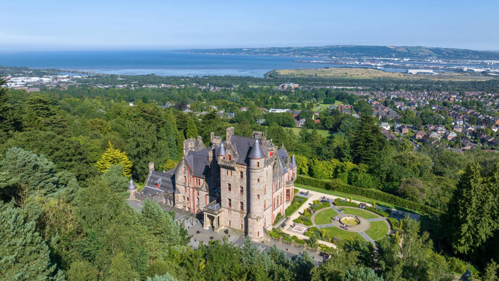 Atemberaubender Blick aus der Luft auf Belfast Castle, umgeben von Wäldern, mit Belfast Lough und der Skyline der Stadt, die sich in der Ferne darunter erstreckt.
