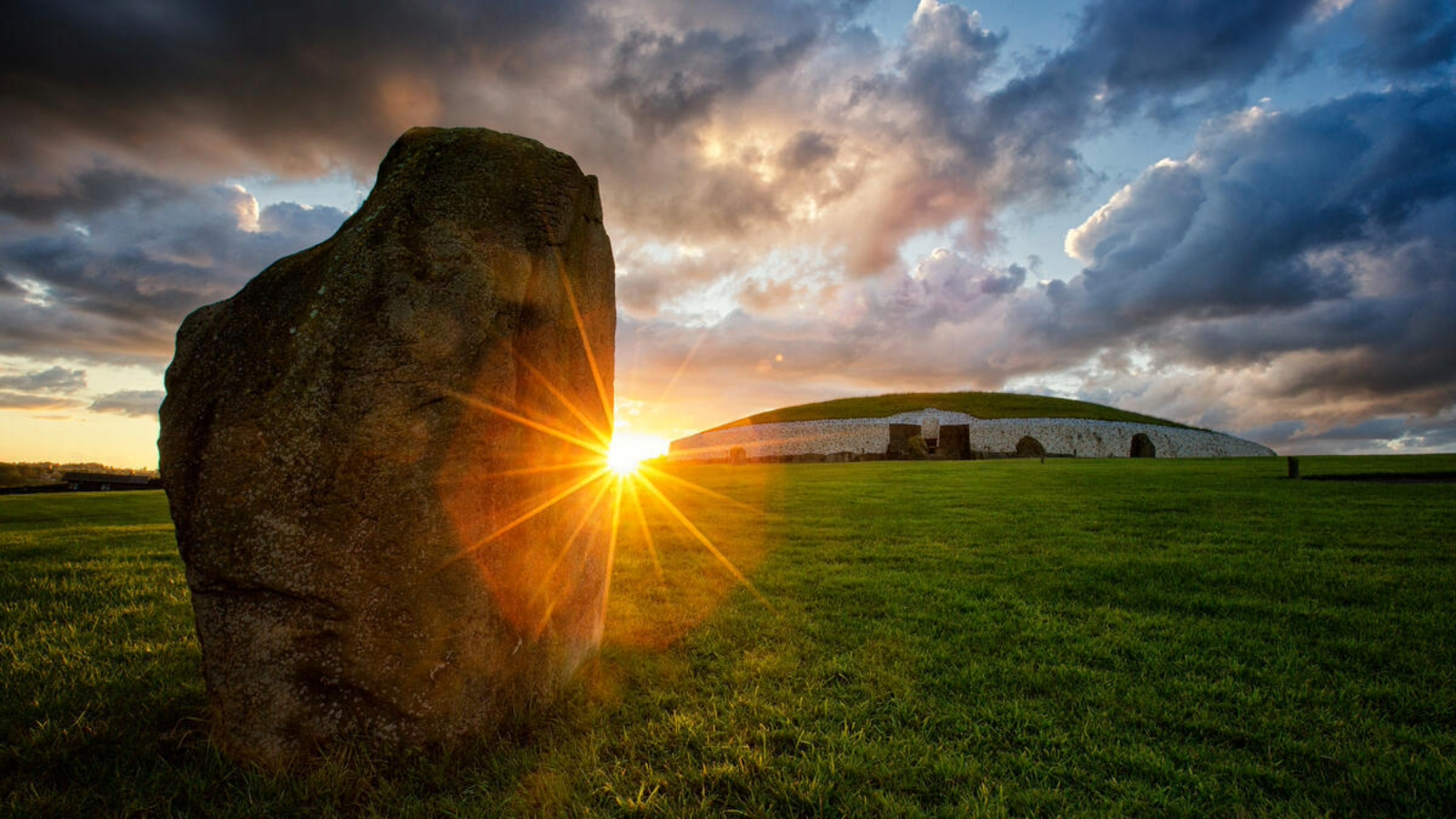 Standing stone at sunrise facing Newgrange passage tomb, County Meath, beneath dramatic clouds.