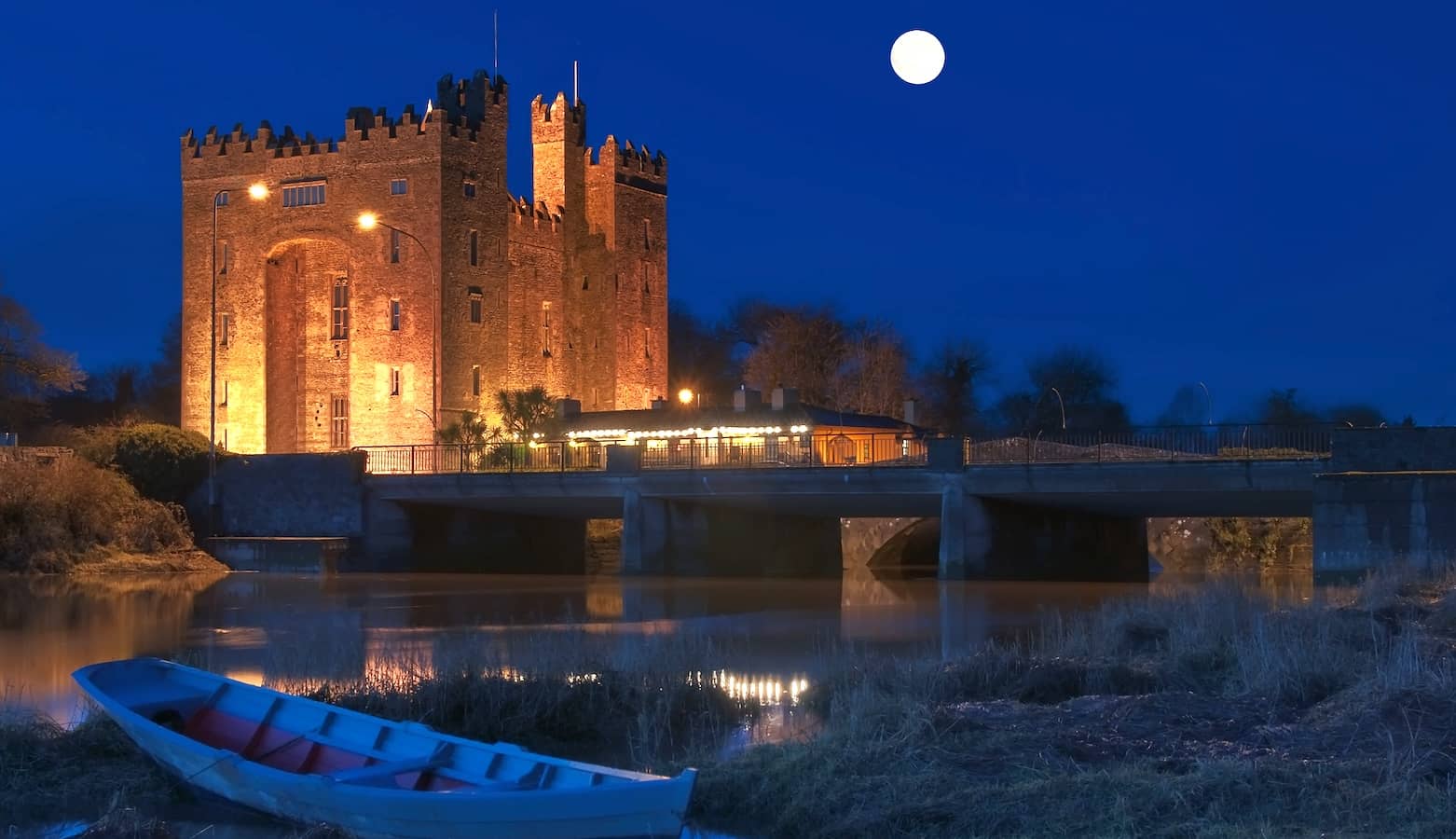 Bunratty Castle lit up at night beneath a full moon, reflected in the calm river beside it.