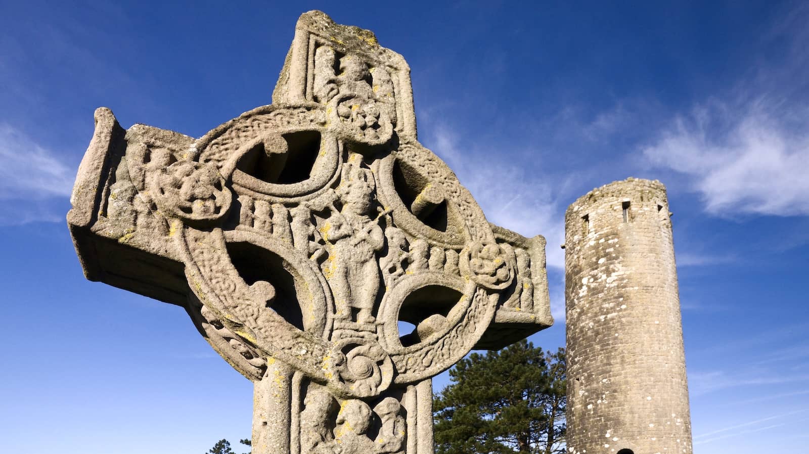 Close-up van een gebeeldhouwd hoog kruis in Clonmacnoise, met op de achtergrond een ronde toren, in county Offaly.