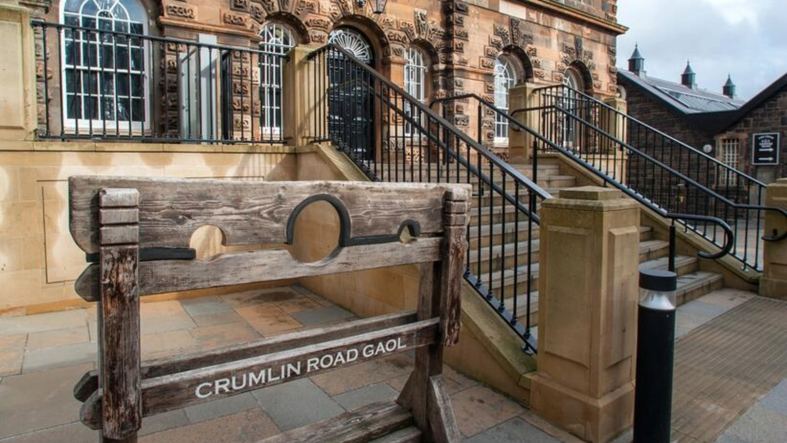 Crumlin Road Gaol entrance in Belfast, with a wooden stocks display and stone steps leading to the doorway.