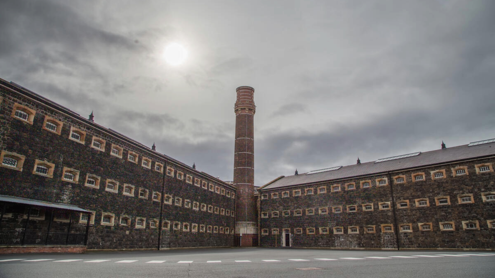 Exterior courtyard of Crumlin Road Gaol, Belfast, with stone walls, small windows and a tall brick chimney.
