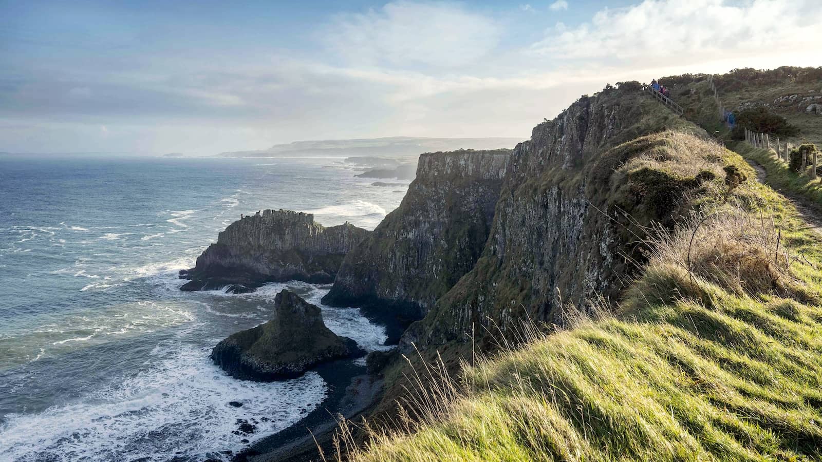 Walkers on a grassy clifftop path overlooking dramatic sea cliffs and waves along the Causeway Coast.