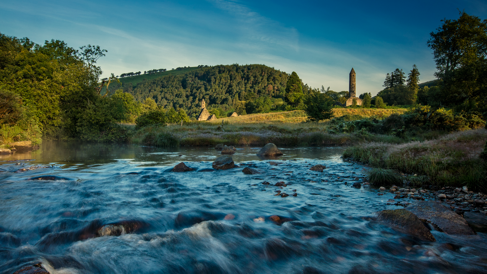 River flowing through Glendalough valley with round tower and monastic ruins beyond.