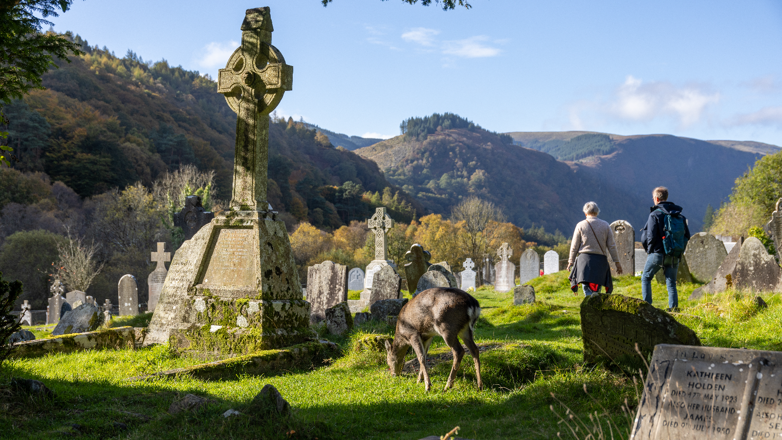 Celtic cross and gravestones in Glendalough cemetery with Wicklow hills in the background.