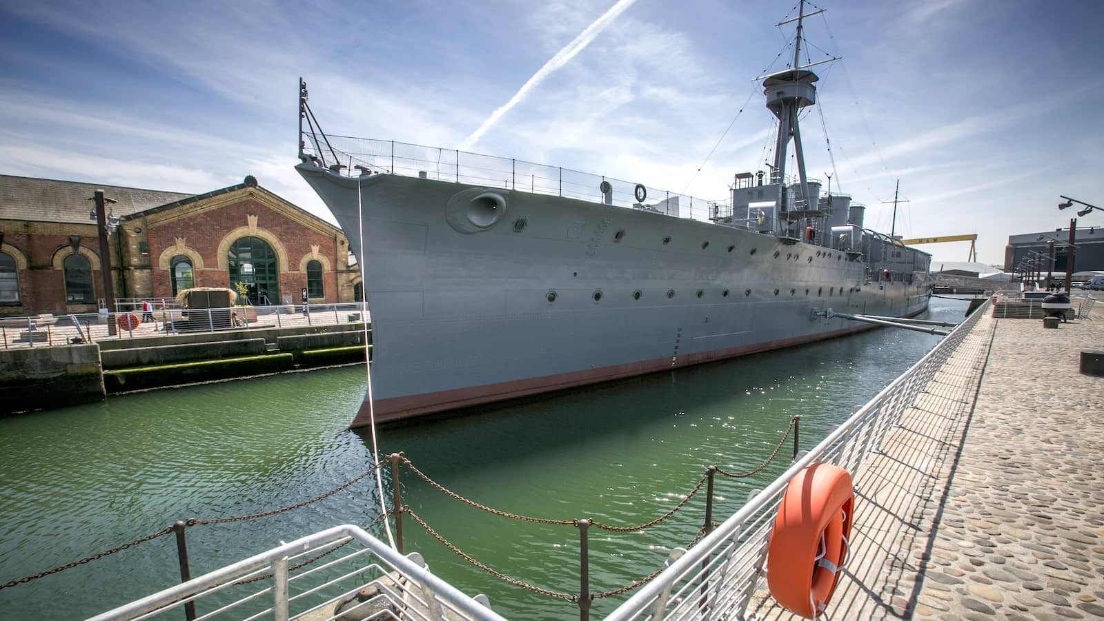 Die HMS Caroline, vor Anker im Titanic Quarter von Belfast und umgeben von historischen Backsteingebäuden sowie der Hafenpromenade.