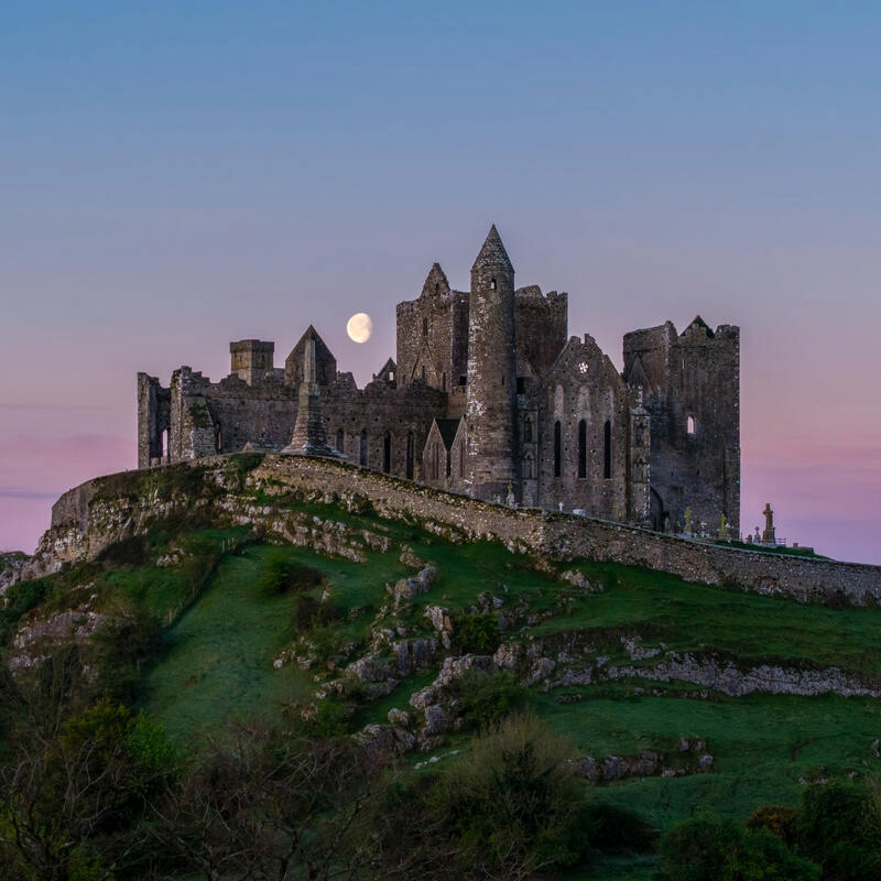 The Rock of Cashel at dusk, with a full moon rising behind the medieval ruins on a green hill in County Tipperary.