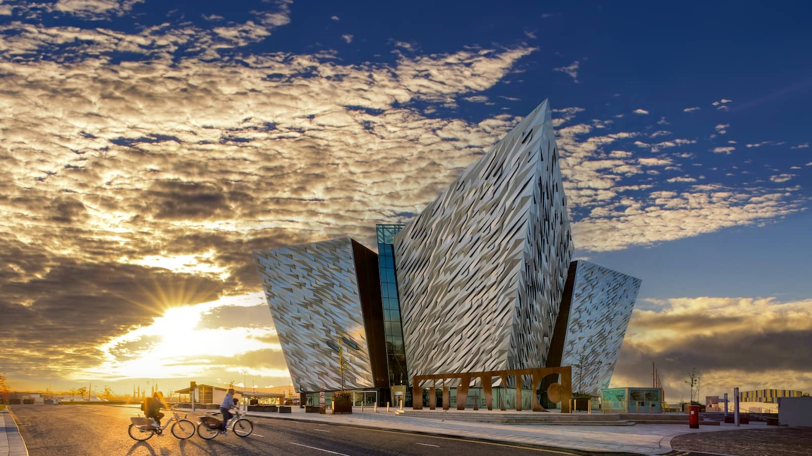Radfahrer, die im Sonnenuntergang am Titanic Belfast vorbeifahren, wobei das goldene Licht die imposante Fassade des Gebäudes erstrahlen lässt.