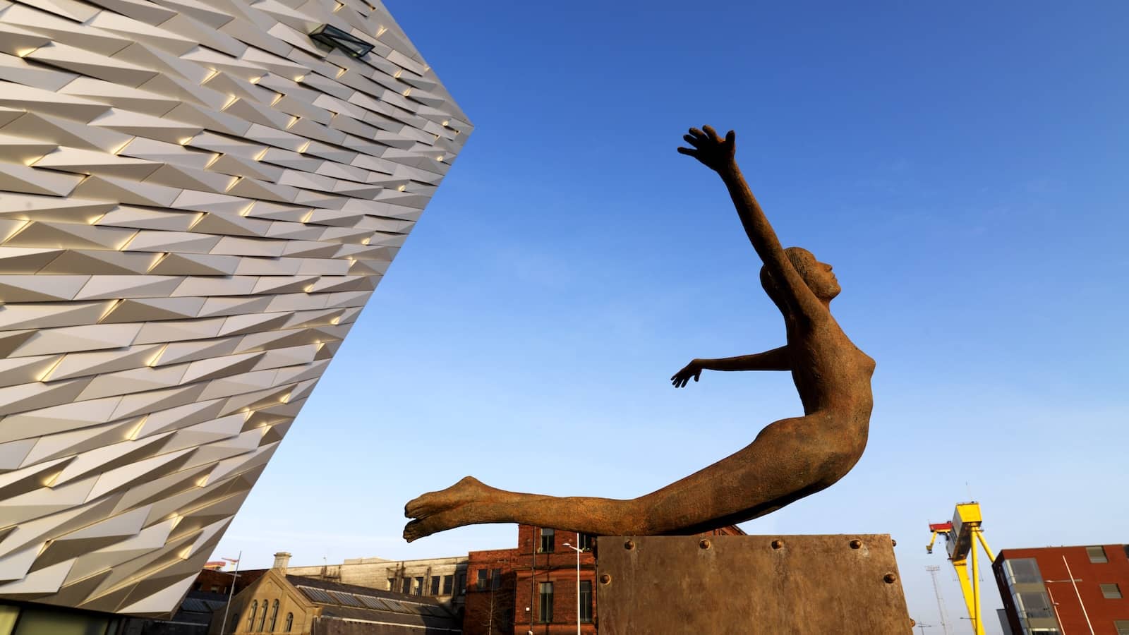 Rostskulptur einer ins Wasser tauchenden Figur neben der eckigen Fassade des Titanic Belfast unter einem klaren blauen Himmel.