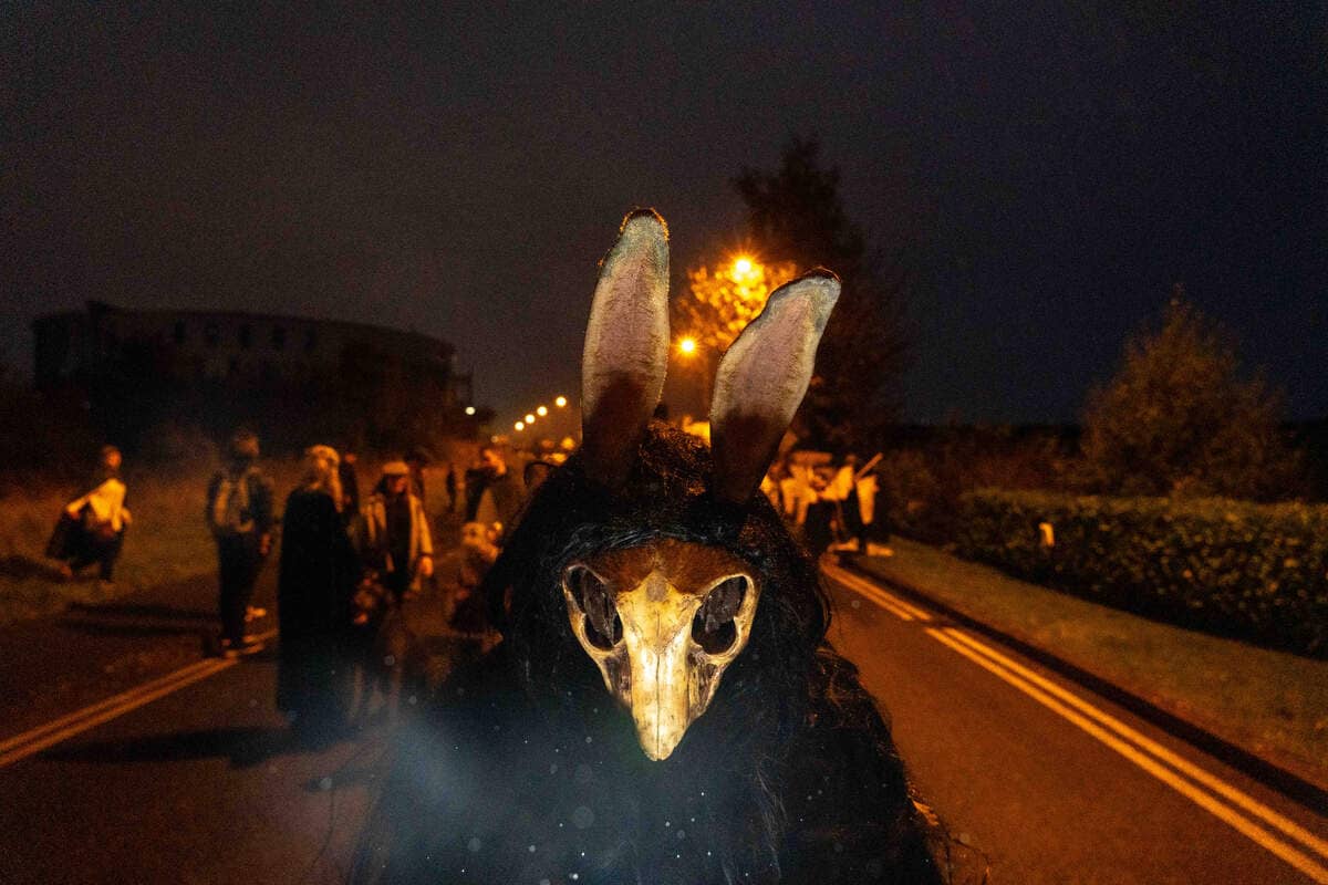 Person in a dark cloak wearing a horned animal skull mask with tall ears, standing on a road at night during the Púca Festival.
