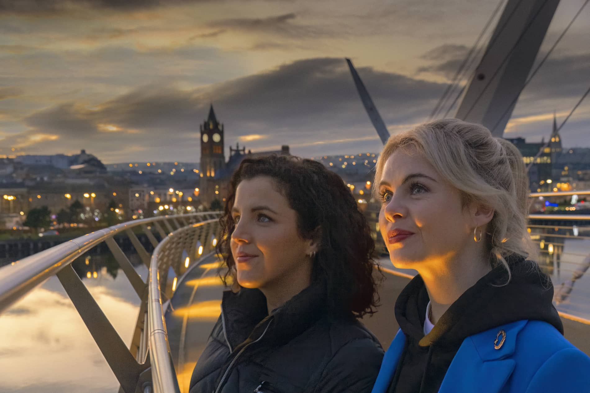 Zwei Frauen bewundern bei Einbruch der Dunkelheit die Stadtlandschaft von der Peace Bridge in Derry~Londonderry aus. Im Hintergrund leuchten die Guildhall und die Lichter der Stadt.