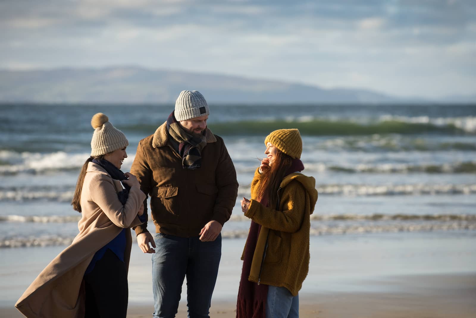 Friends wrapped in winter layers chatting and walking along a breezy Portrush Strand, County Antrim, with waves behind them.