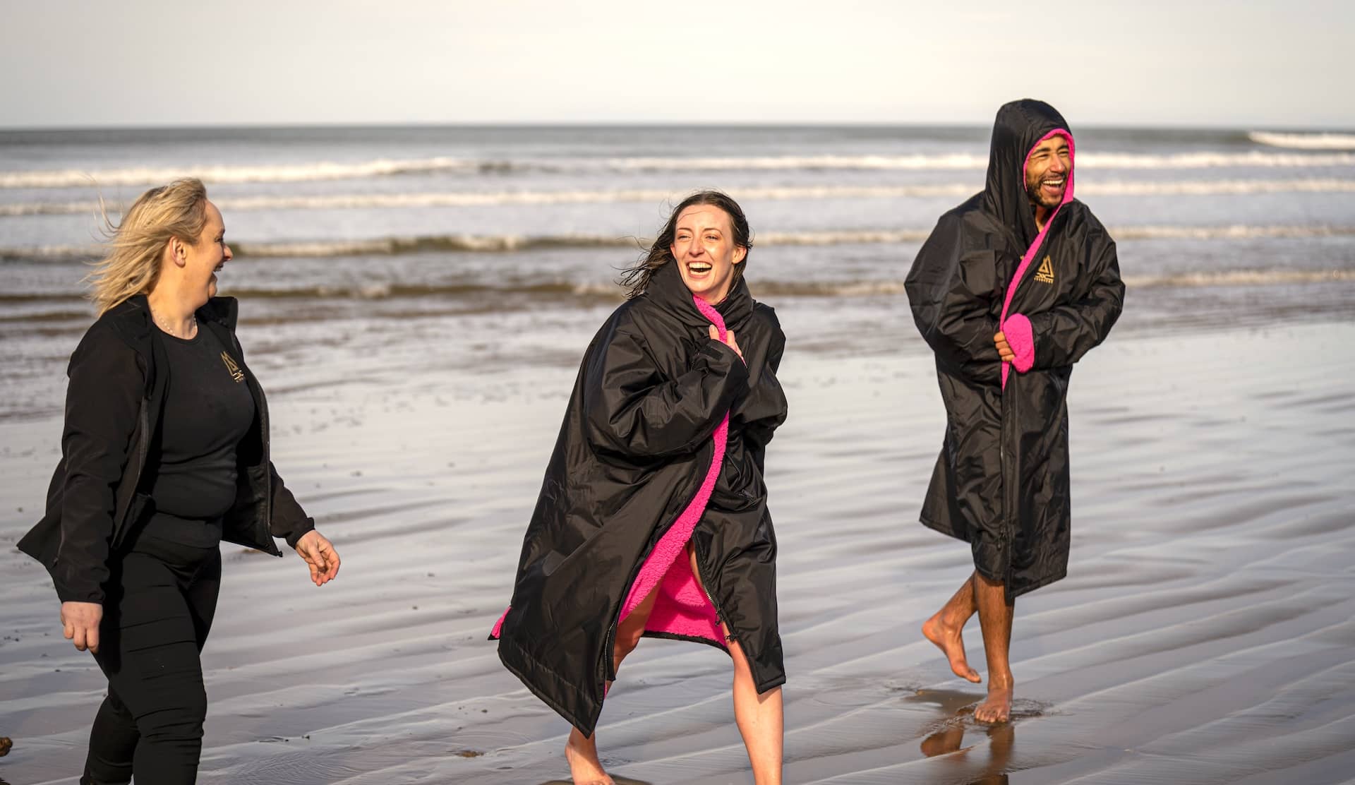 Amigos envueltos en cómodas batas de baño riéndose juntos en una playa después de un baño en el mar con Mussenden Unwind en el condado de Londonderry, con suaves olas de fondo.