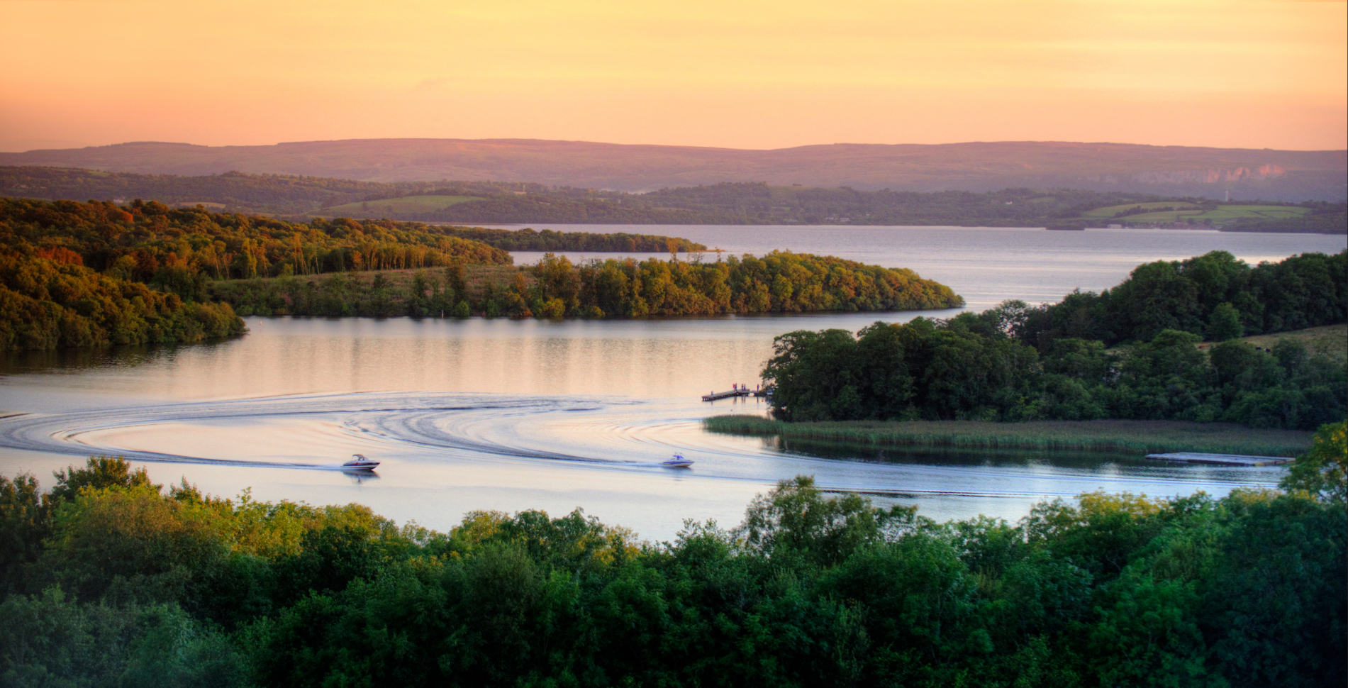 Motorboats create sweeping wakes across Lough Erne at sunset, surrounded by wooded islands in County Fermanagh.