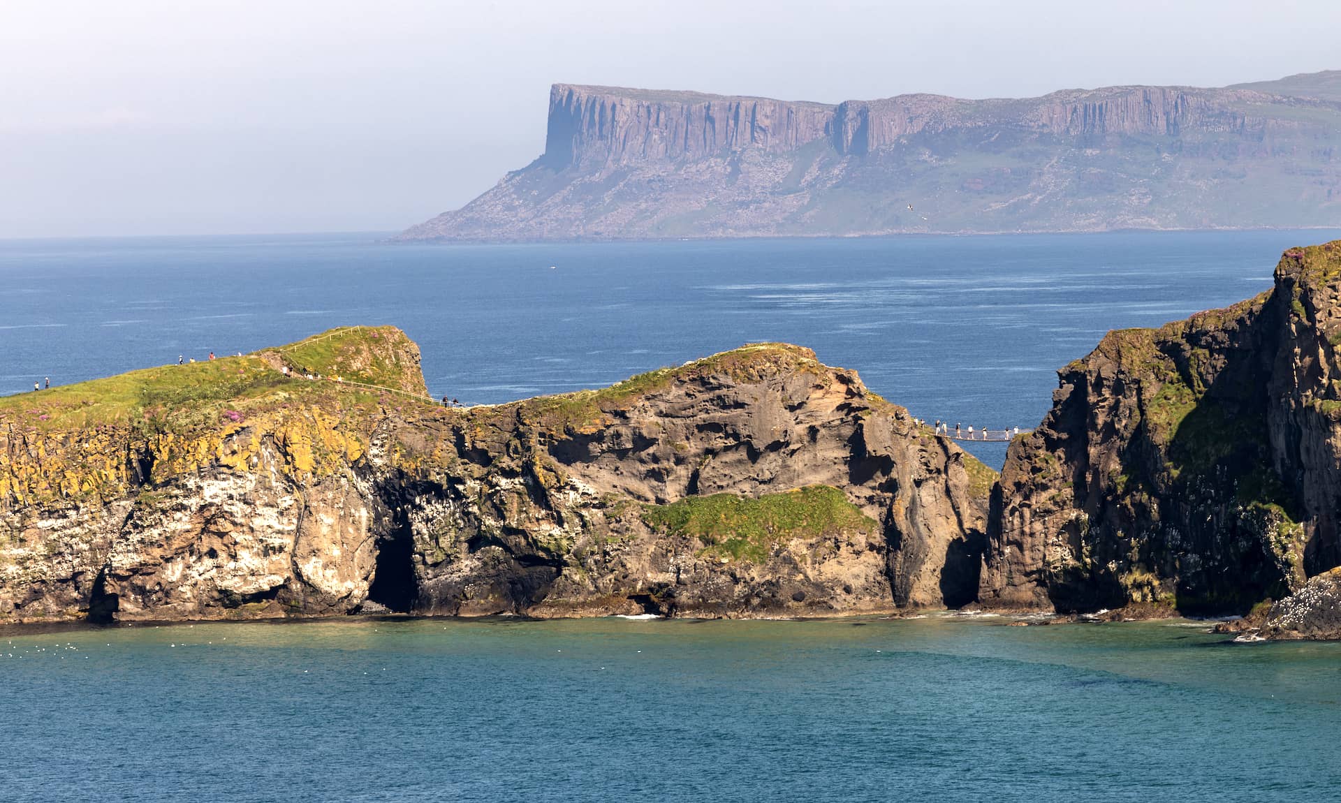 Clifftop views of Carrick-a-Rede with the rope bridge visible above turquoise water and distant headlands in County Antrim.