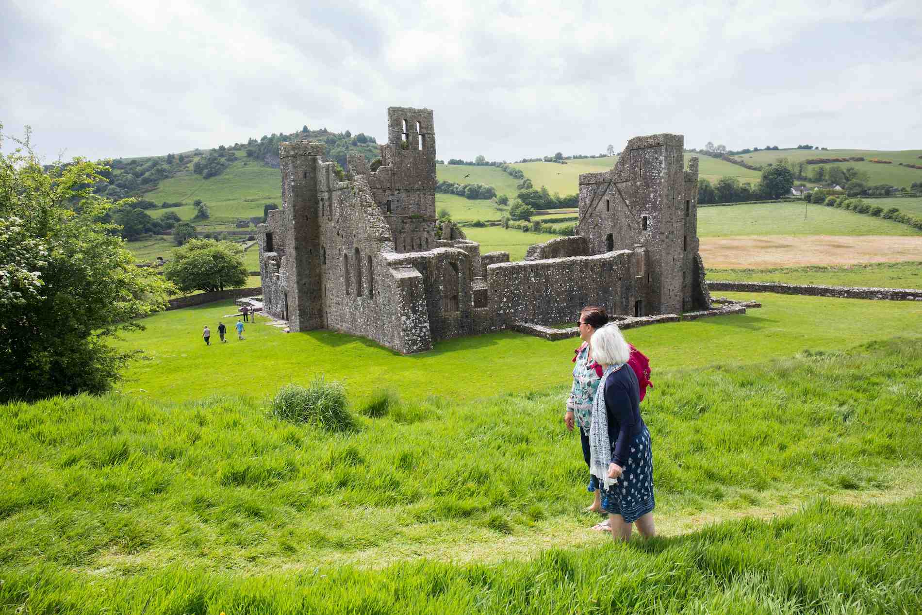 Visitors walking on grassy slopes overlooking the stone ruins of Fore Abbey.