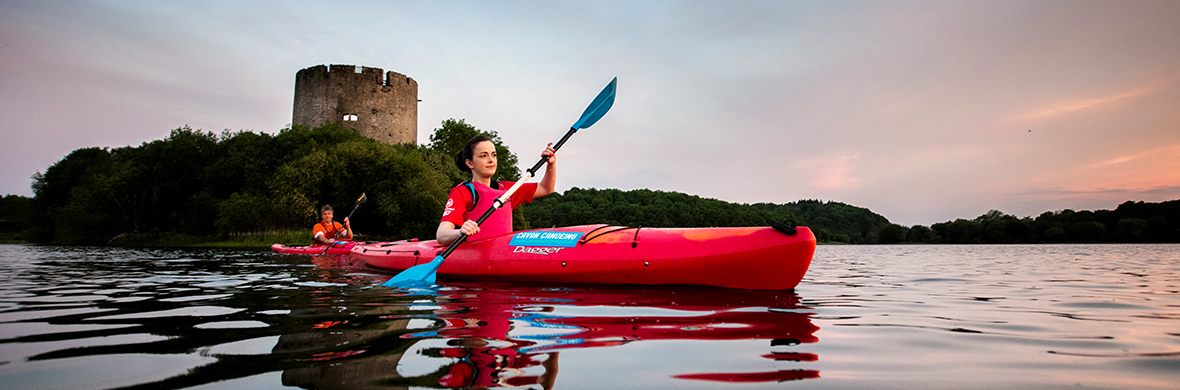 Two people kayaking past Clough Oughter Castle ruins on Lough Oughter in County Cavan at sunset.