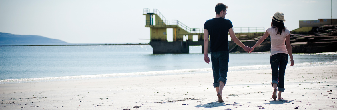 Couple walking hand in hand on Salthill Beach with the diving platform in the distance.