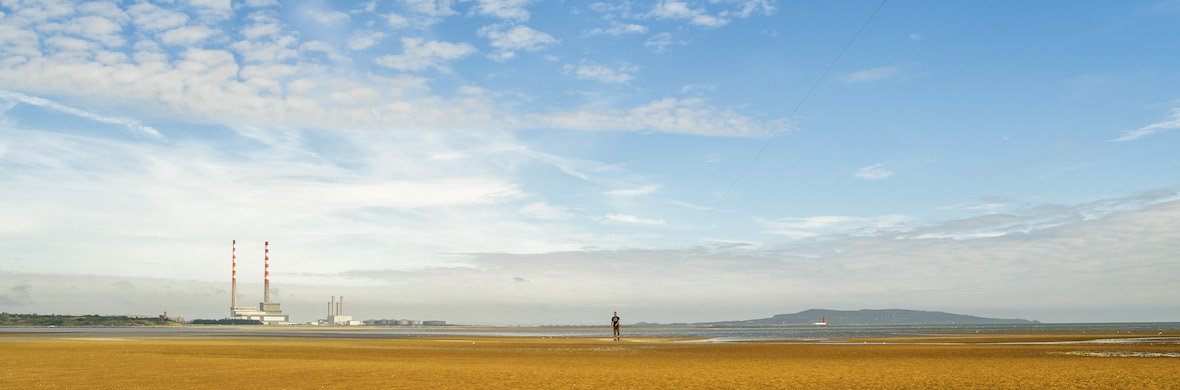 Person walks on Sandymount Strand with views of Poolbeg chimneys and Howth across Dublin Bay under a blue sky.