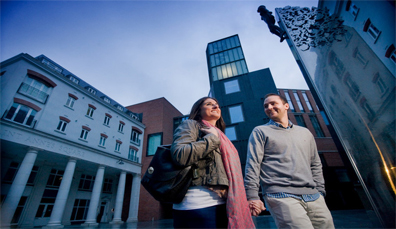 Coppia che si tiene per mano fuori dal MAC a Belfast al crepuscolo con edifici moderni sullo sfondo.