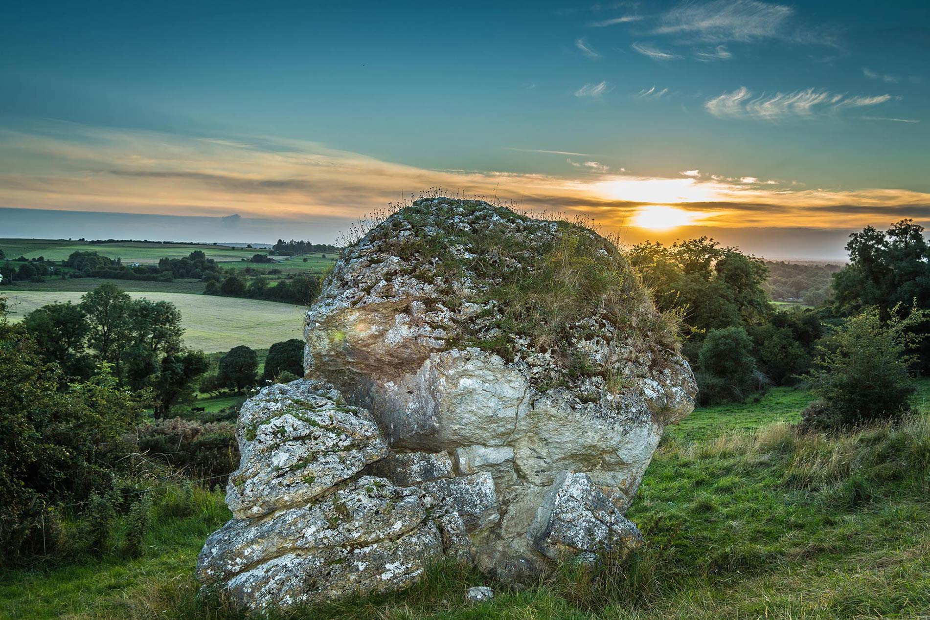 Oude rotsblok op de Hill of Uisneach met glooiende groene velden en een gouden zonsondergang in de verte.