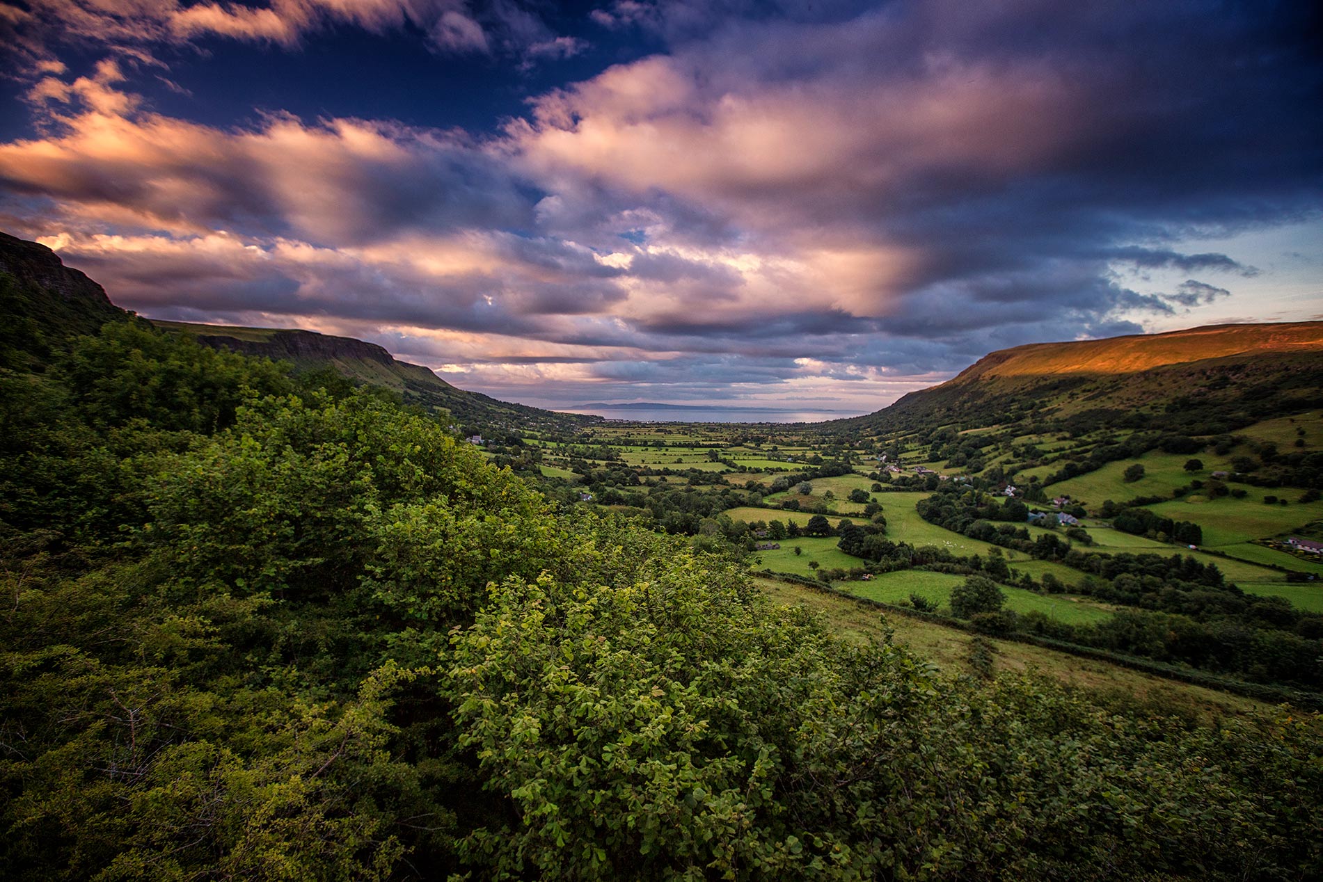Weelderig dal in Glenariff Glen omgeven door beboste heuvels, met een levendige zonsondergang die de horizon verlicht.