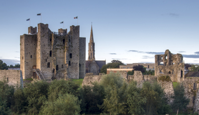 Ruins of Trim Castle with fluttering flags and a tall church spire behind, viewed from across treetops at dusk.