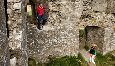 Two visitors explore the stone ruins of the Rock of Dunamase, with grass underfoot and worn masonry walls.