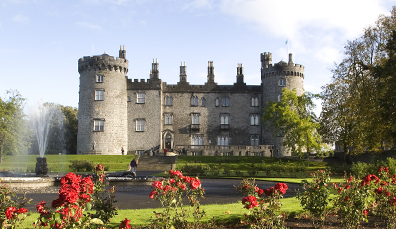 Kilkenny Castle with symmetrical towers and a manicured rose garden in front, framed by a fountain and bright sky.