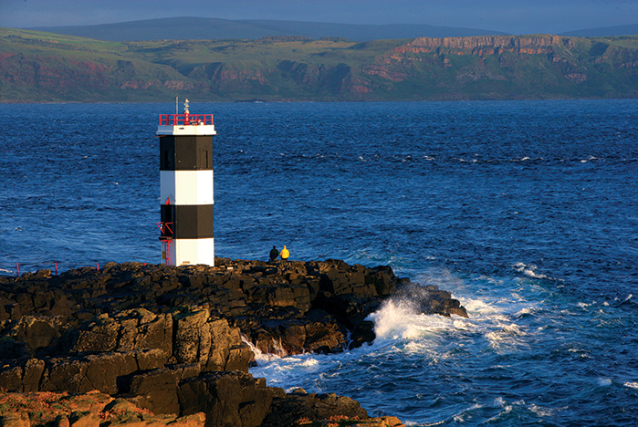 El faro de Rue Point, de rayas en blanco y negro, se eleva de las oscuras rocas costeras mientras las olas chocan contra la orilla.